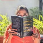 a woman sitting holding best book about travel to read in her hands in clay color dress the book title shows journeys beyond and within it is a green cover and in the background you can see plants