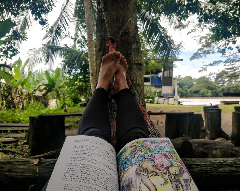 best travel books to read in the photo author is lying down with book on her lap in malaysia kinabatagan river homestay