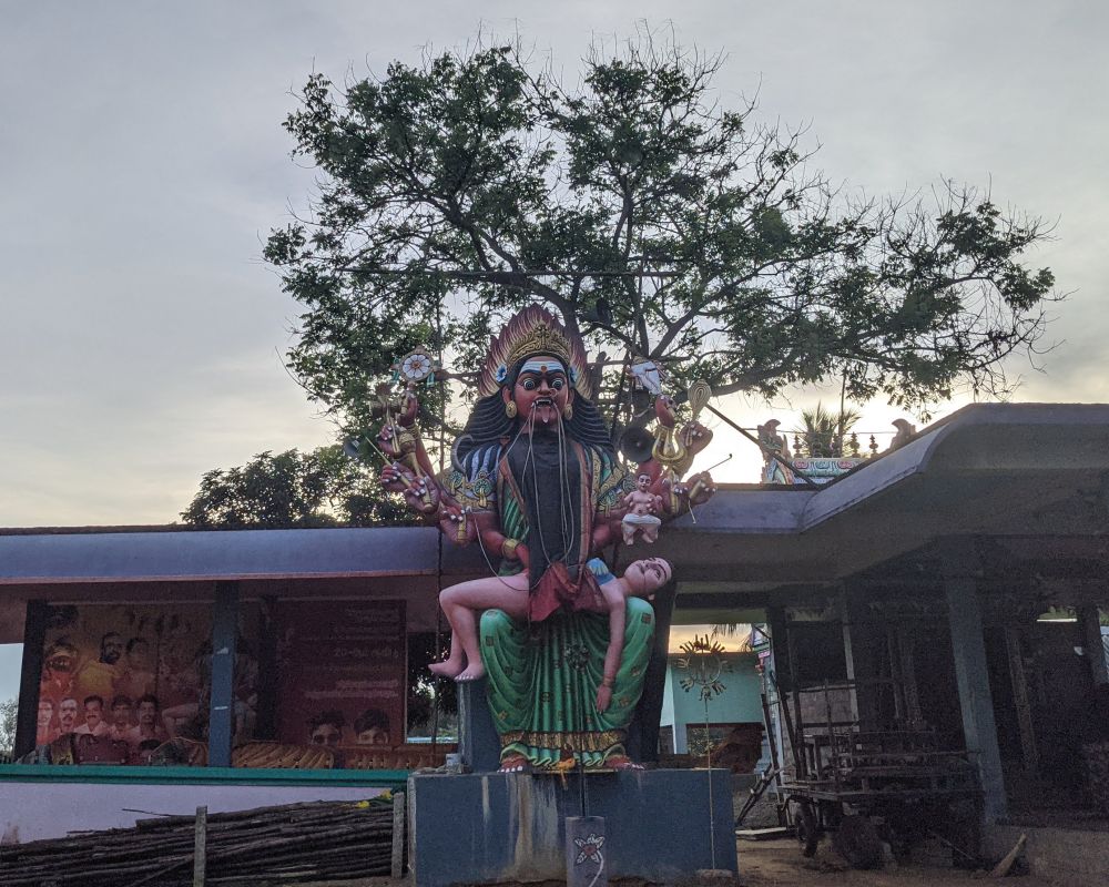 the statue of a goddess with a dead person she killed in her lap.  atree behind the statue, tamil nadu