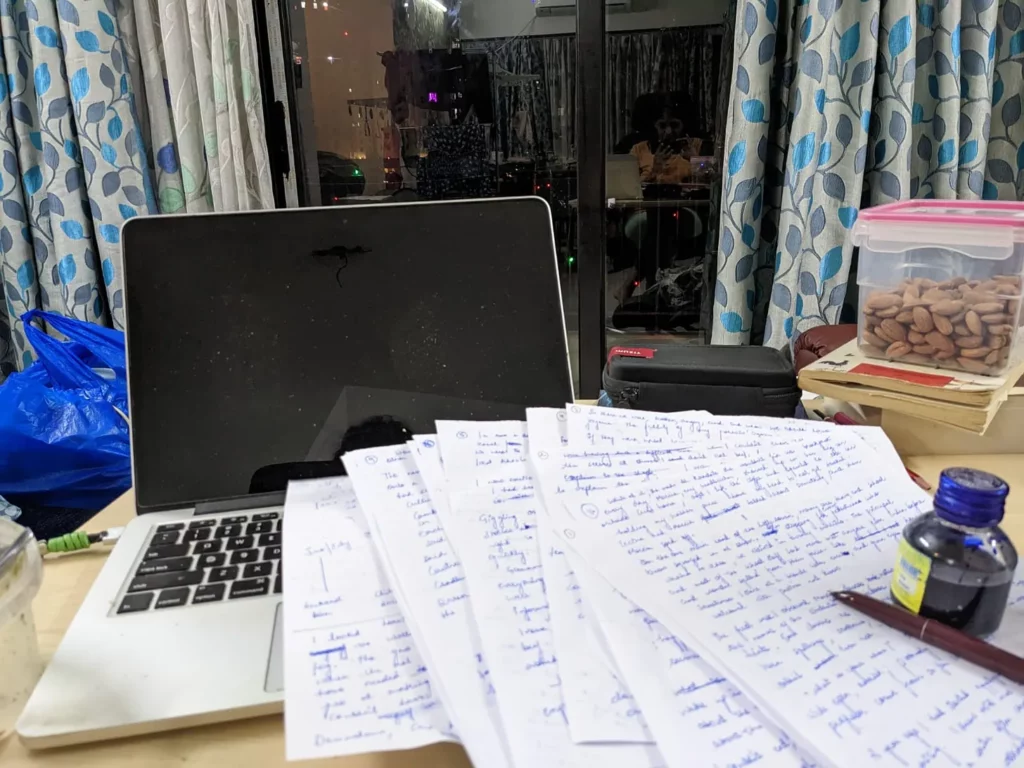 writing in my friends home with pen and paper. laptop, filled sheets with english writing in blue ink, ink pot, almonds, and a glass window beyond with the author visible in it.