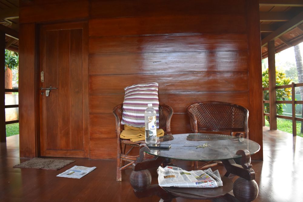 a wooden porch showing a glass-top table and two chairs. Behind the table-chairs, a room whose gate is closed can be seen. surrounding the porch is a garden.