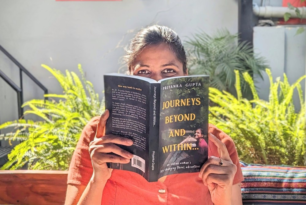 a woman sitting holding a great travel book to read in her hands in clay color dress the book title shows journeys beyond and within it is a green cover and in the background you can see plants