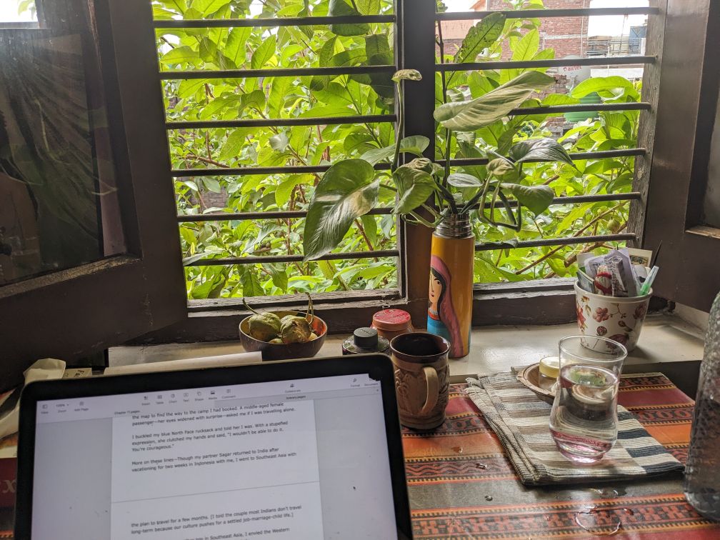 a computer on a table and a window beyond with a green guava tree outside and cups, ink pot, a colorful vase etc on the table too shown in the article for write a first draft of book