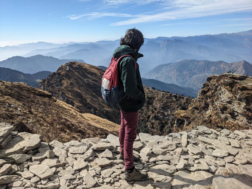 a man standing with a colorful bag on a mountain on rocks with blue mountains rolling around him. open blue sky